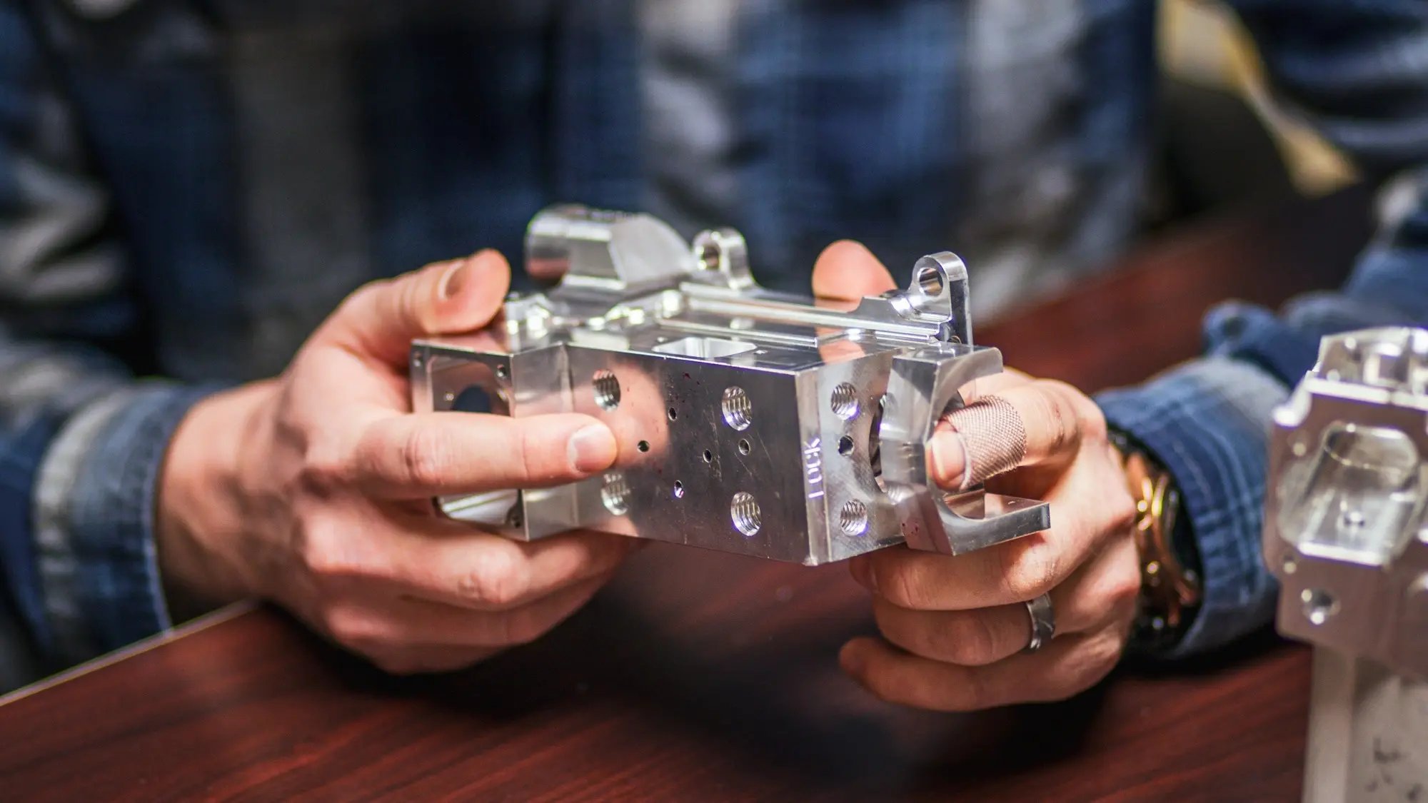 A person in a denim jacket holds a precision-machined aluminum block with threaded holes and mechanical fittings