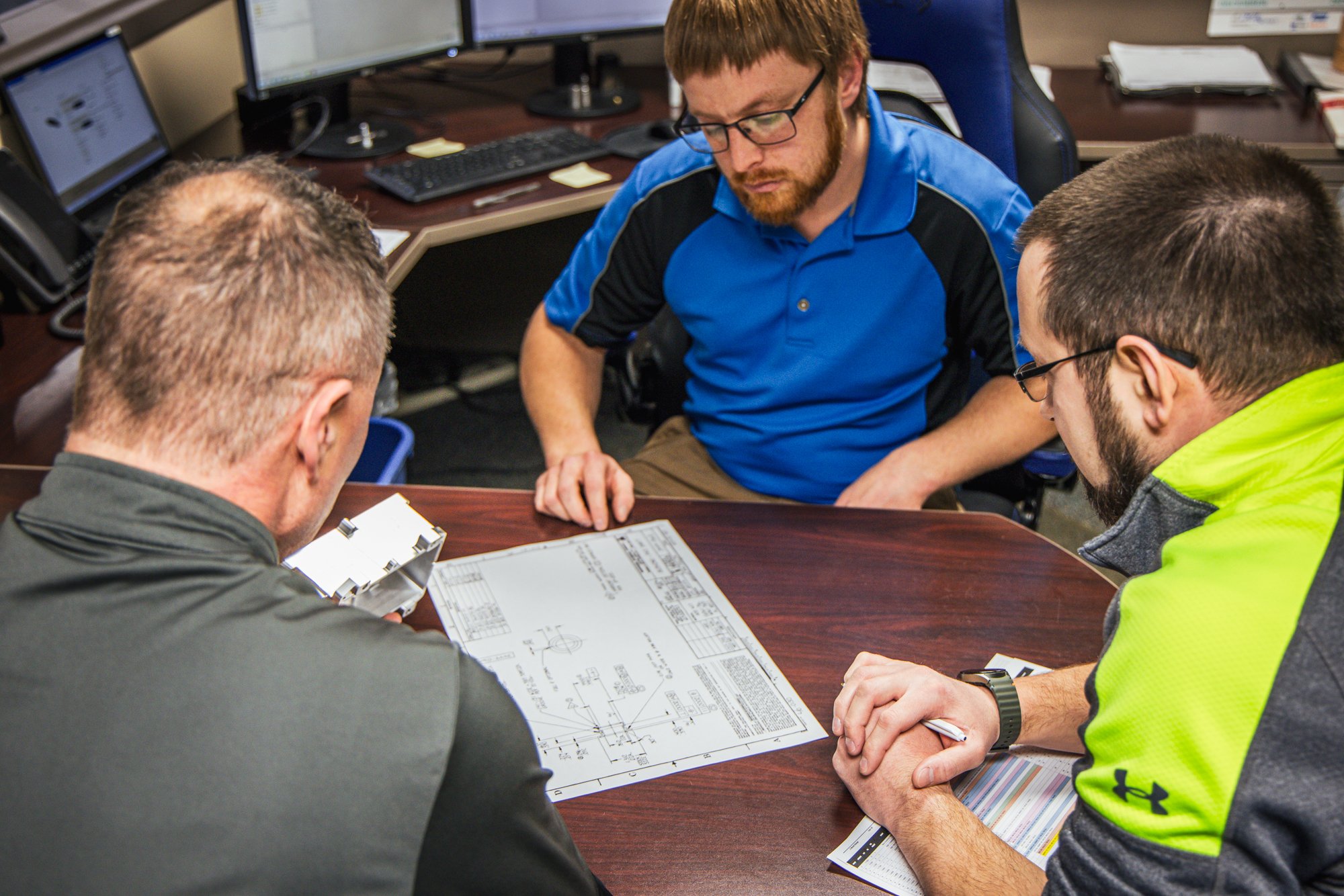 Three engineers reviewing a technical drawing and machined part at a conference table.