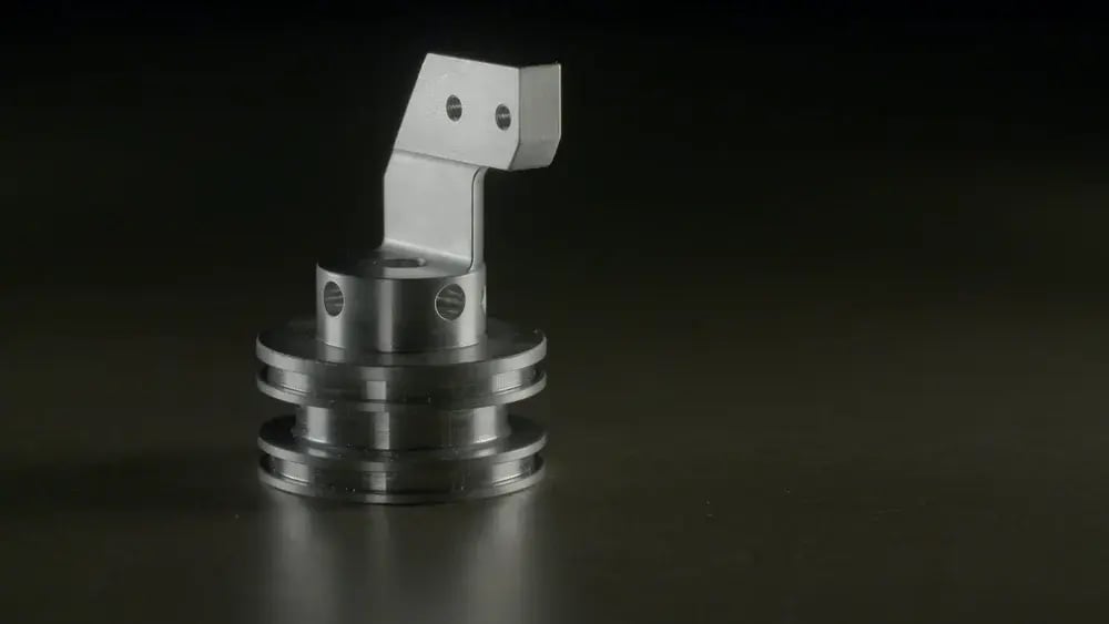 A complex precision machined metal component with a flanged base and angled bracket on a dark surface.