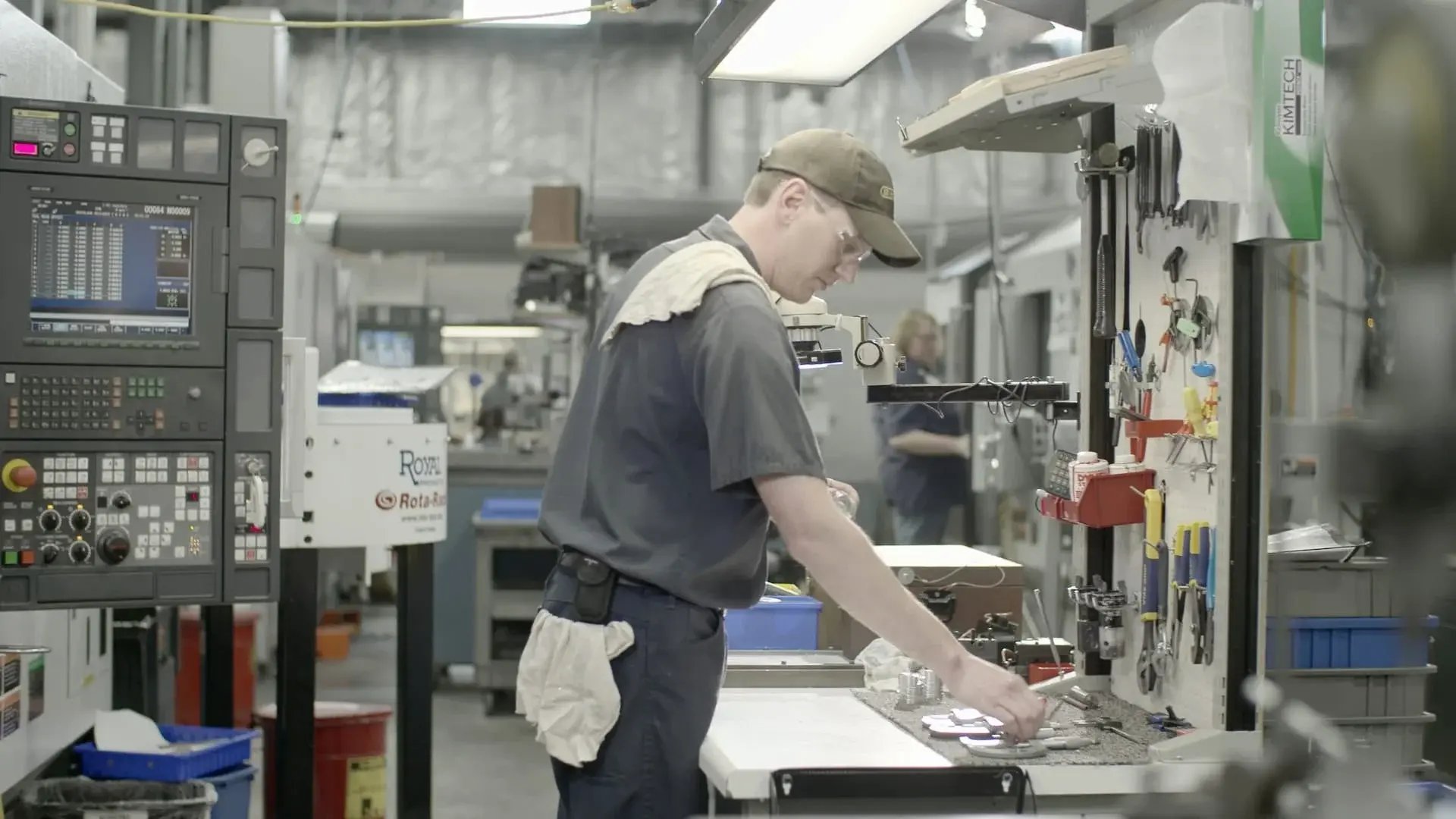A machinist working at a tool station next to a CNC control panel on a busy production floor.